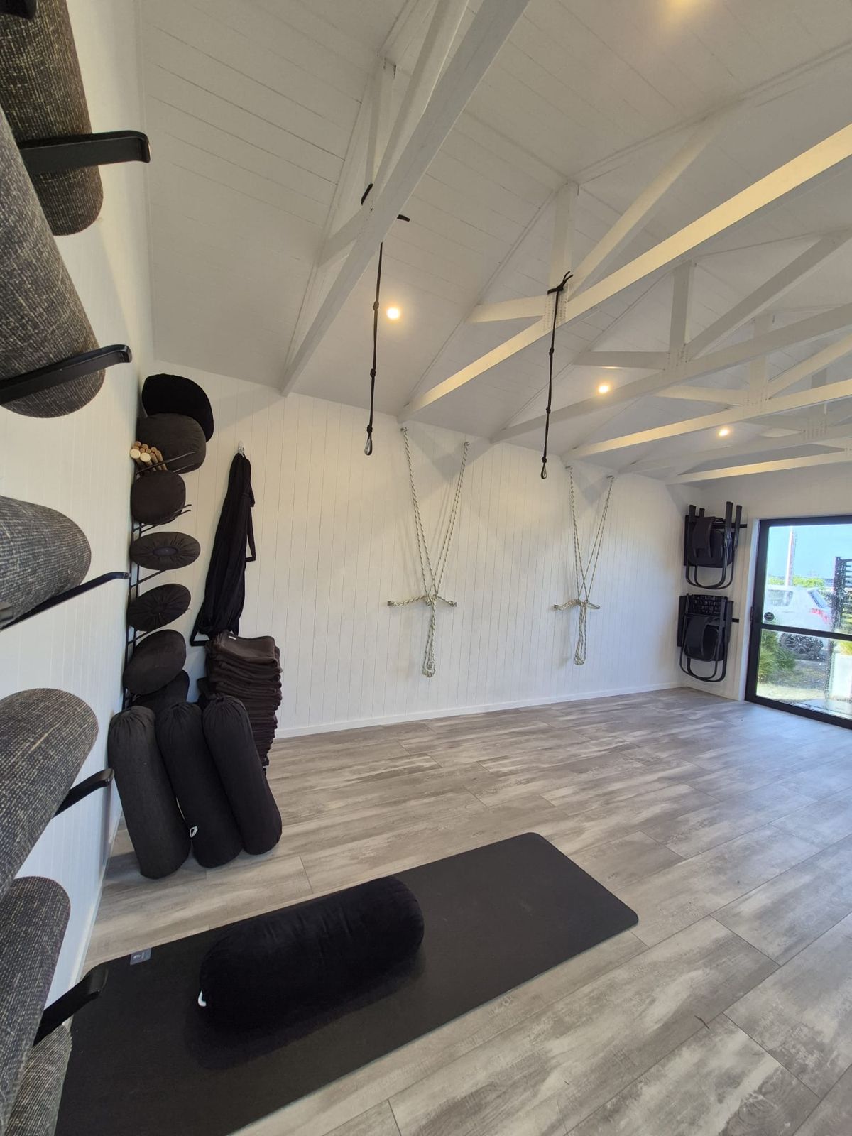 The Yoga Boutique studio: white timber walls and trusses, wall ropes and ceiling ropes, a rack of bolsters, round cushions, blankets and folding chairs, with a mat laid out in front